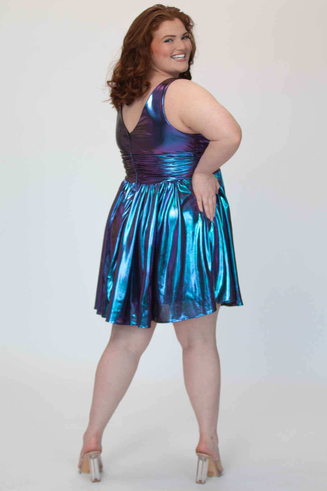 A-line flowy short party dress, SC8156 by Sydney's Closet. Back view. In the color Prism, a  blue/purple metallic knit fabric. Thick bra-friendly straps, pleated v-neckline bodice and waist band, long invisible center back zipper. V- back. Available in Prism, Peacock and Raven black, sizes 14-32. 
Model in picture is 5'9. 