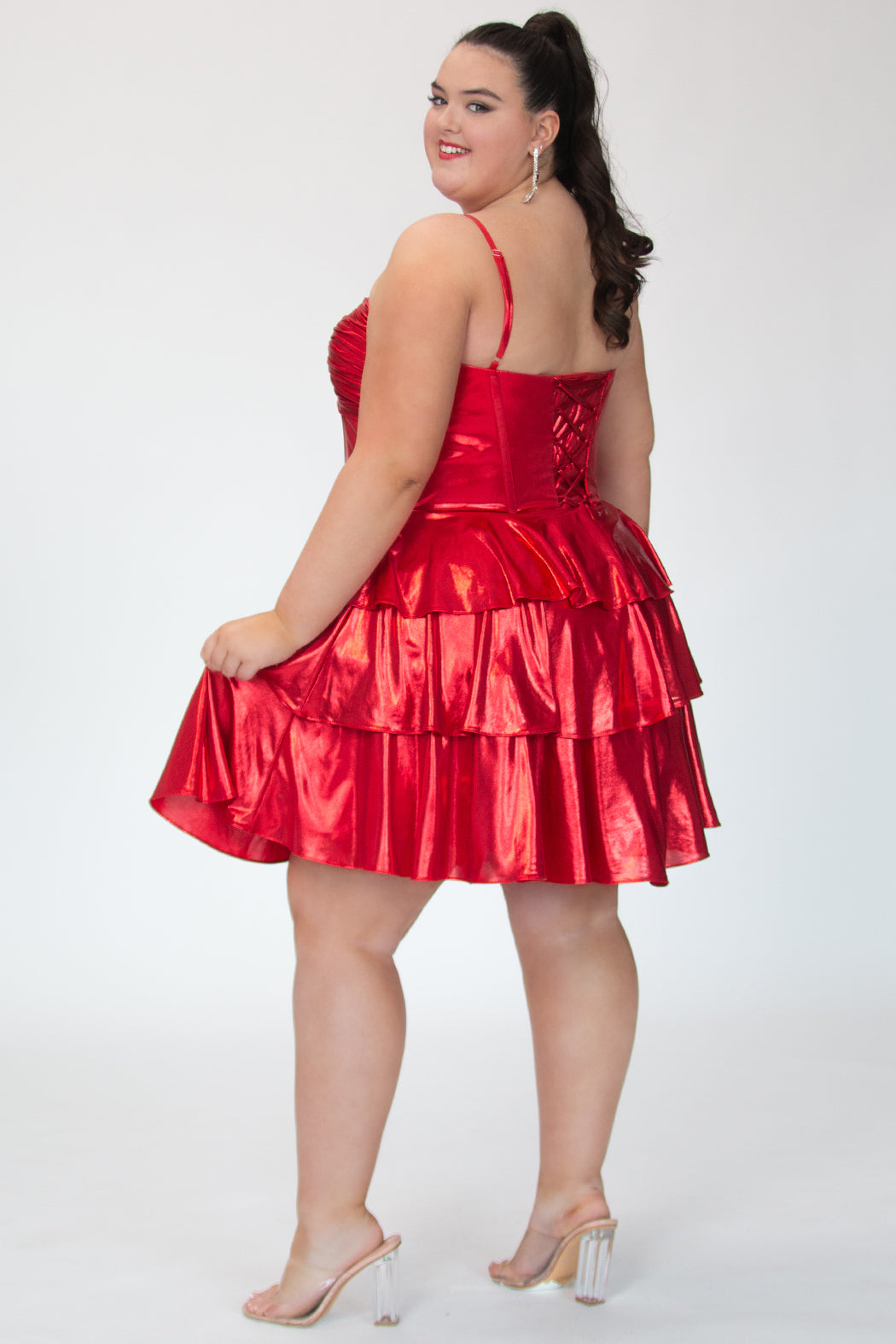 SC8161, metallic Ruby Red flowy plus size party dress by Sydney's Closet. Back view. Ruched knotted bodice with spaghetti straps, exposed boning and lace up back with modesty panel. A-line tiered skirt. Metallic stretch knit and knit lining. Available in Ruby red and Gold, sizes 14-24. 