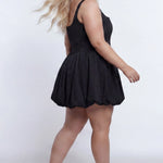 Plus-size model wearing a black sleeveless taffeta mini dress with a square neckline, structured corset-inspired bodice with exposed boning, and voluminous bubble hem skirt. She is styled with long flowing blonde hair, drop earrings, and nude platform ankle-strap heels, walking to the side against a light gray studio background. Back view.

Sydney's Closet SC8166 in black