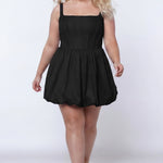 Plus-size model wearing a black sleeveless taffeta mini dress with a square neckline, structured corset-inspired bodice with exposed boning, and voluminous bubble hem skirt. She is styled with long flowing blonde hair, drop earrings, and nude platform ankle-strap heels, walking toward the camera against a light gray studio background. Front view.

Sydney's Closet SC8166 in black