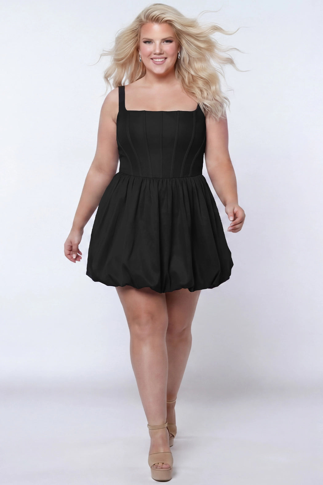 Plus-size model wearing a black sleeveless taffeta mini dress with a square neckline, structured corset-inspired bodice with exposed boning, and voluminous bubble hem skirt. She is styled with long flowing blonde hair, drop earrings, and nude platform ankle-strap heels, walking toward the camera against a light gray studio background. Front view.

Sydney's Closet SC8166 in black