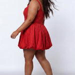 Plus-size model wearing a red sleeveless taffeta mini dress with a square neckline, structured corset-inspired bodice with exposed boning, and voluminous bubble hem skirt. She is styled with long flowing brown hair, drop earrings, and nude platform ankle-strap heels, walking to the side against a light gray studio background. Back view.

Sydney's Closet SC8166 in red