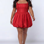 Plus-size model wearing a red sleeveless taffeta mini dress with a square neckline, structured corset-inspired bodice with exposed boning, and voluminous bubble hem skirt. She is styled with long flowing brown hair, drop earrings, and nude platform ankle-strap heels, walking toward the camera against a light gray studio background. Front view.

Sydney's Closet SC8166 in red
