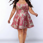 Plus-size model wearing a sleeveless a-line mini dress with a scoop neckline and metallic floral brocade pattern in fuchsia pink, gold, and champagne tones. The dress features straps, a fitted bodice and a flared skirt with soft pleats. She is styled with long flowing dark brown hair, dangling earrings, and nude platform ankle-strap heels, walking toward the camera against a light gray studio background.

Sydney's Closet SC8169 in fuchsia pink floral