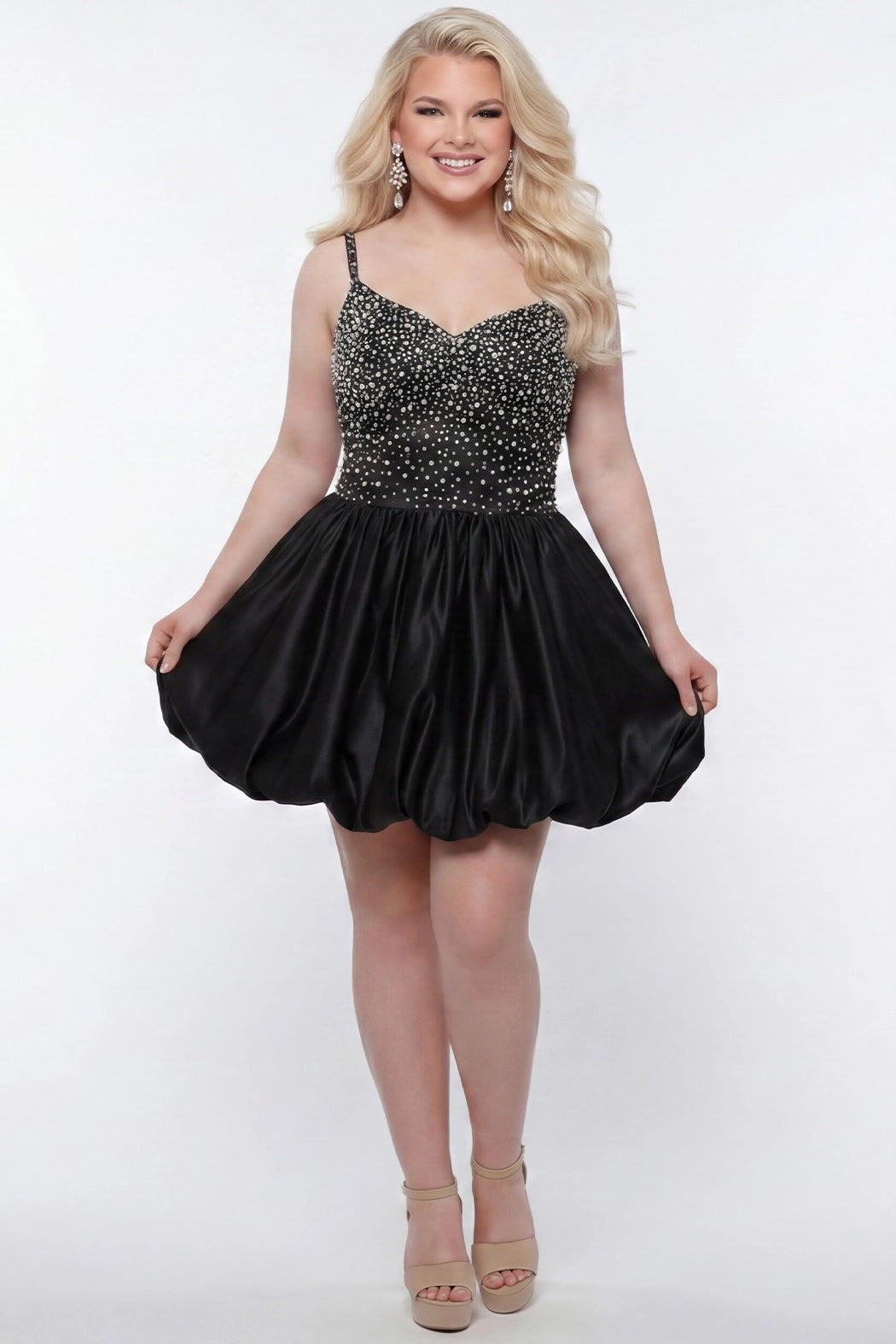 Plus-size model wearing a black sleeveless mini dress with a V-neckline, rhinestone & pearl embellished fitted bodice, and a voluminous bubble hem skirt. She is styled with long wavy blonde hair, dangling crystal earrings, and nude platform ankle-strap heels, posing against a light gray studio background while holding the sides of the skirt.

Sydney's Closet SC8171 in black