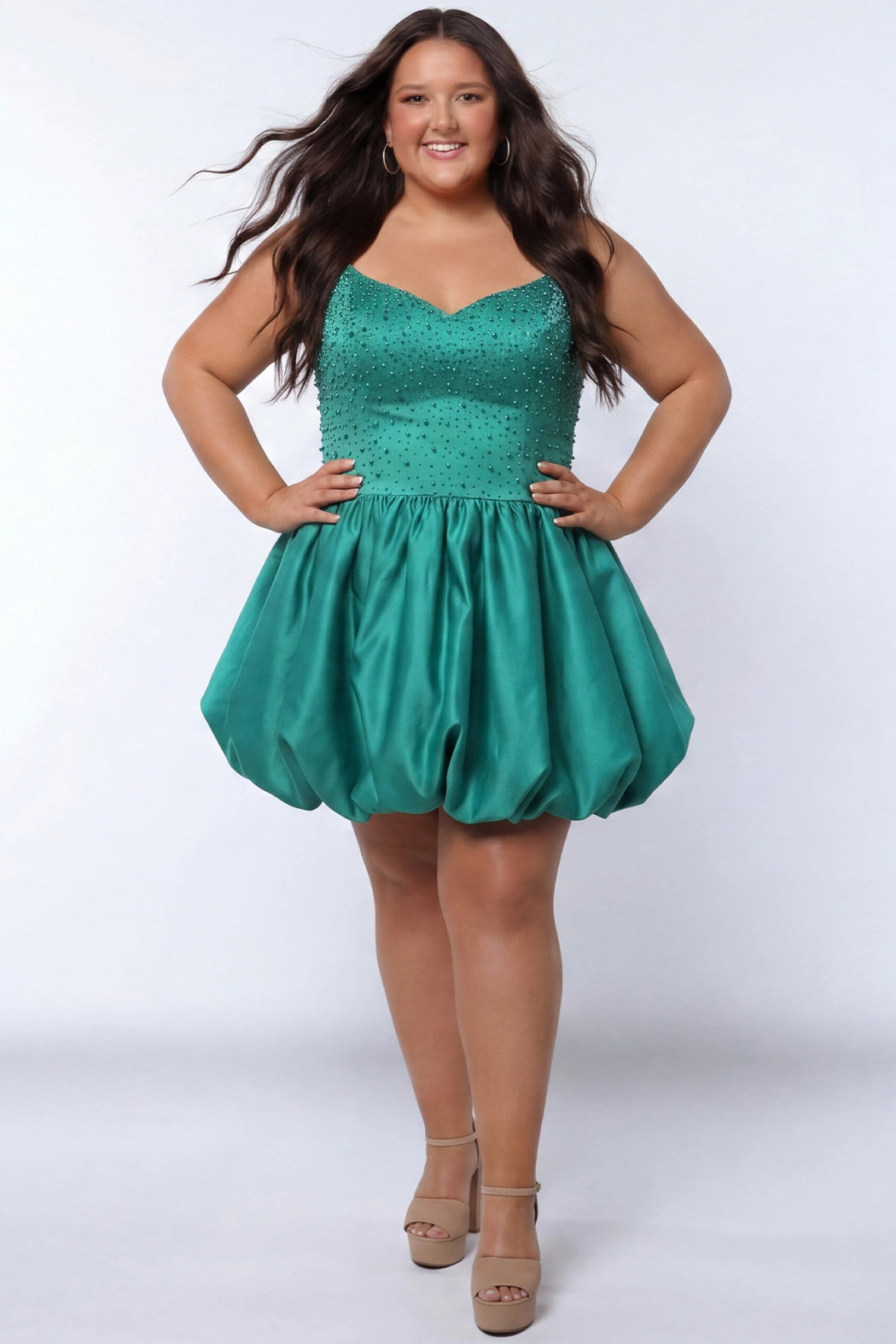 Plus-size model wearing a emerald green sleeveless mini dress with a V-neckline, rhinestone & pearl embellished fitted bodice, and a voluminous bubble hem skirt. She is styled with long wavy brown hair, dangling crystal earrings, and nude platform ankle-strap heels, posing against a light gray studio background while her hands are on her hips.

Sydney's Closet SC8171 in green