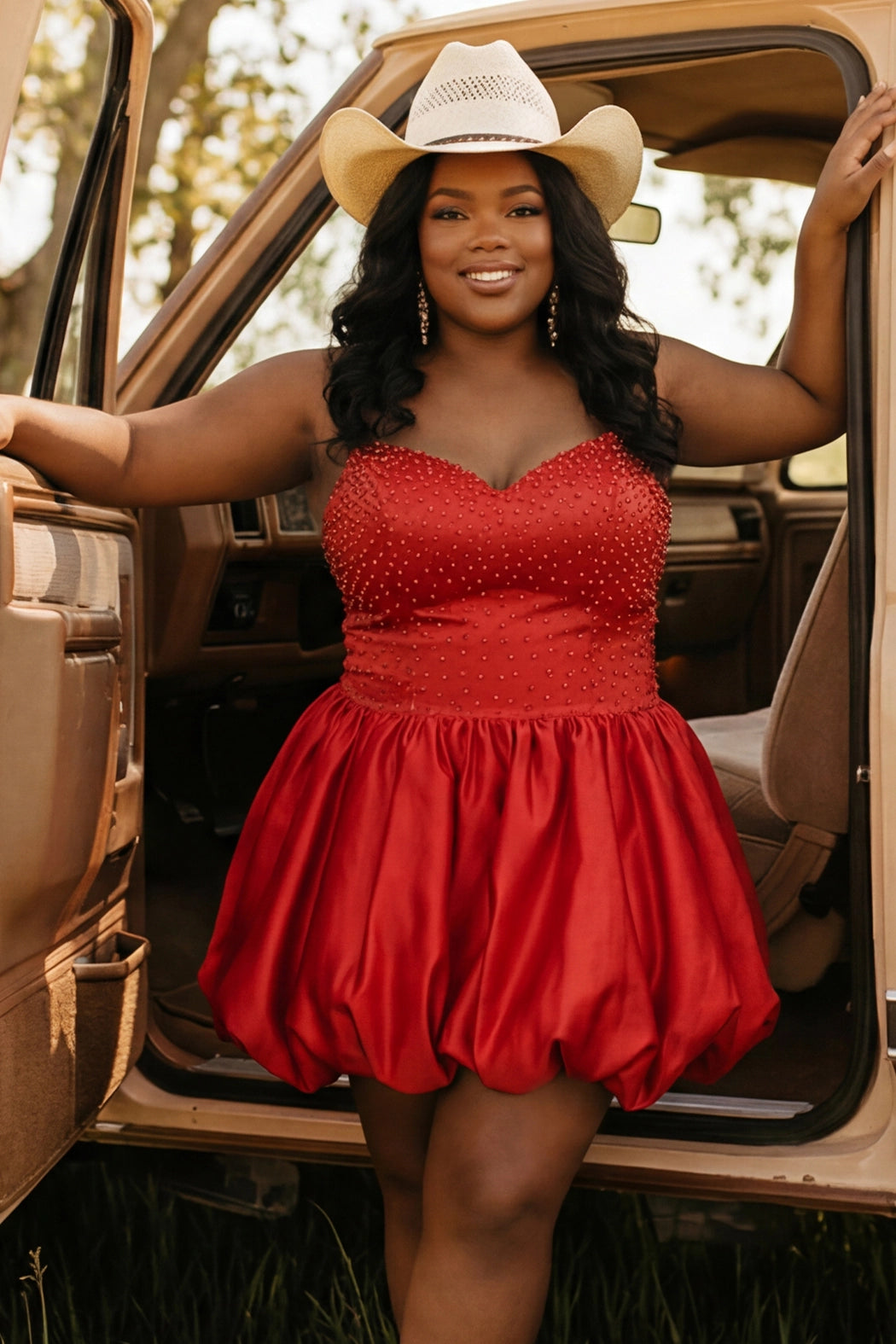 Plus-size model wearing a red spaghetti-strap mini dress with a sweetheart neckline, fitted rhinestone-embellished bodice, and voluminous bubble hem skirt. She is styled with a white cowboy hat, long wavy black hair, and dangling earrings, posing in the open doorway of a vintage tan truck. The outdoor setting features warm natural light and a rustic country-inspired aesthetic.

Sydney's Closet SC8171 in red