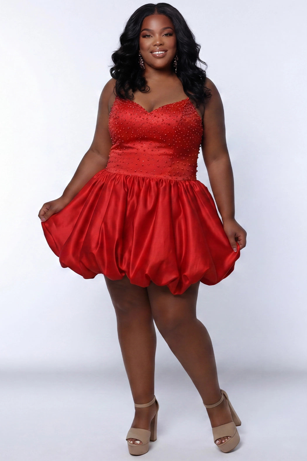 Plus-size model wearing a red sleeveless mini dress with a V-neckline, rhinestone & pearl embellished fitted bodice, and a voluminous bubble hem skirt. She is styled with long wavy black hair, dangling crystal earrings, and nude platform ankle-strap heels, posing against a light gray studio background while holding the sides of the skirt.

Sydney's Closet SC8171 in red