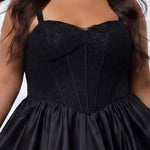 Plus-size model wearing a black sleeveless mini dress with a structured corset-style bodice and sweetheart neckline. The dress features lace detailing on the bodice and a voluminous satin bubble hem skirt. She is styled with long wavy dark brown hair, dangling earrings, and clear open-toe heels, posing against a light gray studio background while holding the sides of the skirt. Detail view. 

Sydney's Closet SC8173