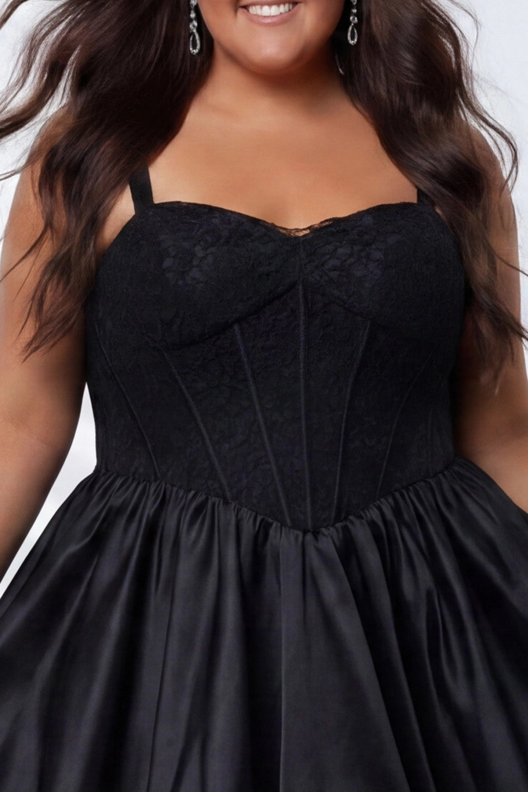 Plus-size model wearing a black sleeveless mini dress with a structured corset-style bodice and sweetheart neckline. The dress features lace detailing on the bodice and a voluminous satin bubble hem skirt. She is styled with long wavy dark brown hair, dangling earrings, and clear open-toe heels, posing against a light gray studio background while holding the sides of the skirt. Detail view. 

Sydney's Closet SC8173