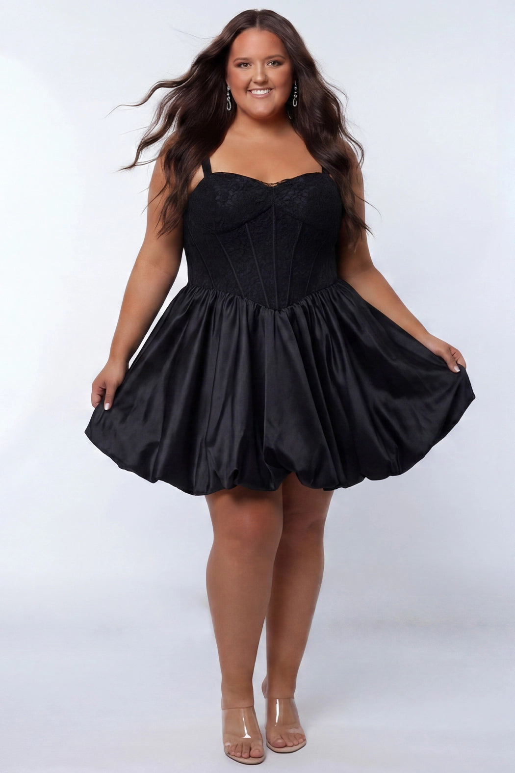 Plus-size model wearing a black sleeveless mini dress with a structured corset-style bodice and sweetheart neckline. The dress features lace detailing on the bodice and a voluminous satin bubble hem skirt. She is styled with long wavy dark brown hair, dangling earrings, and clear open-toe heels, posing against a light gray studio background while holding the sides of the skirt. Front view. 

Sydney's Closet SC8173