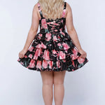 Plus-size model wearing a black and pink floral sleeveless mini dress with layered ruffle skirt. The dress features a square neckline, fitted bodice, and tiered ruffles at the waist and hem. She is styled with long wavy blonde hair, lace-up back with modesty panel, and nude platform ankle-strap heels, posing against a studio background. Back view. 

Sydney's Closet SC8174