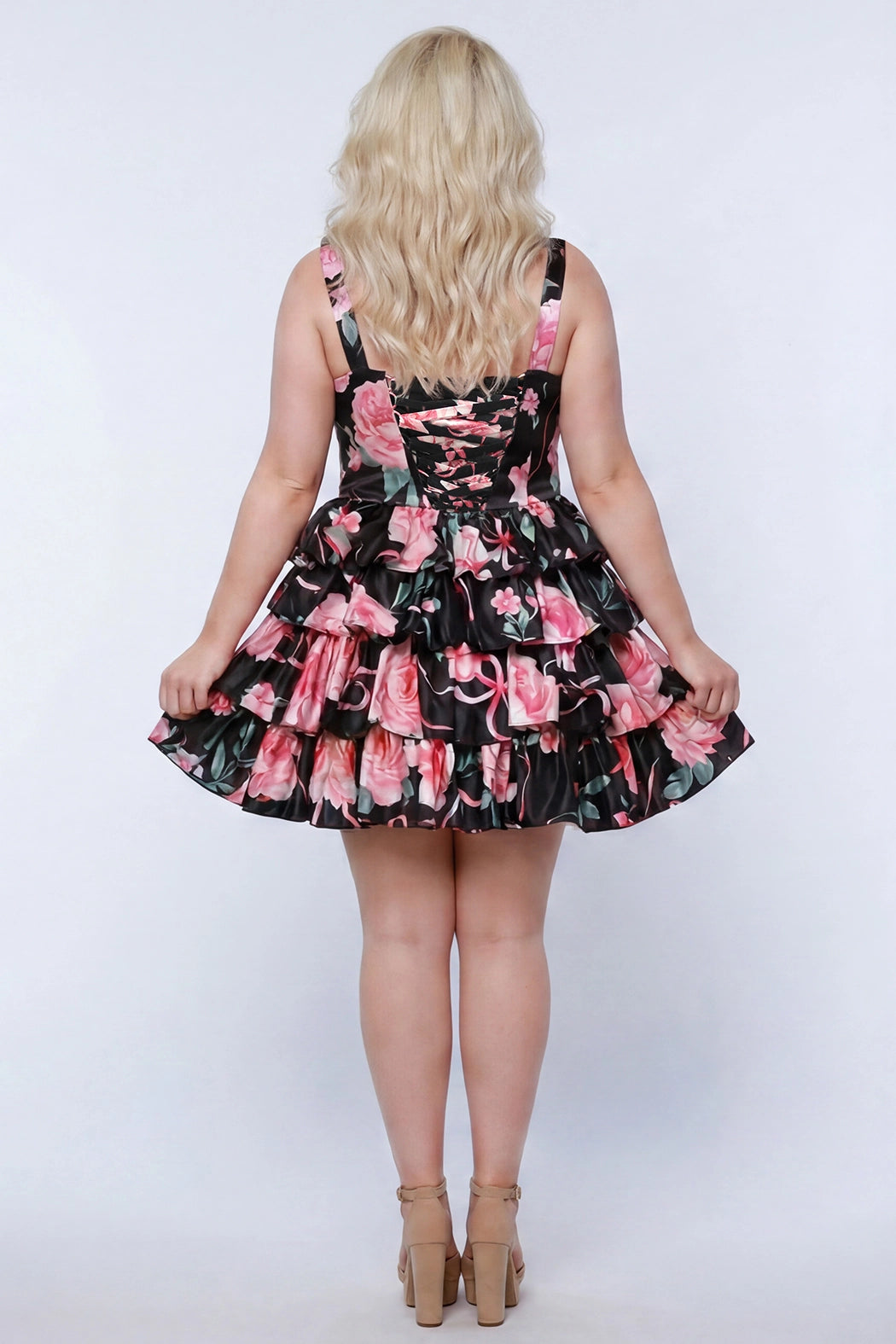 Plus-size model wearing a black and pink floral sleeveless mini dress with layered ruffle skirt. The dress features a square neckline, fitted bodice, and tiered ruffles at the waist and hem. She is styled with long wavy blonde hair, lace-up back with modesty panel, and nude platform ankle-strap heels, posing against a studio background. Back view. 

Sydney's Closet SC8174