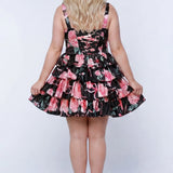 Plus-size model wearing a black and pink floral sleeveless mini dress with layered ruffle skirt. The dress features a square neckline, fitted bodice, and tiered ruffles at the waist and hem. She is styled with long wavy blonde hair, lace-up back with modesty panel, and nude platform ankle-strap heels, posing against a studio background. Back view. 

Sydney's Closet SC8174