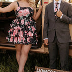 Young couple outdoors beside a vintage tan vehicle. The young woman wears a black and pink sleeveless mini dress with a pink floral print, square neckline, and tiered ruffle skirt, paired with nude platform ankle-strap heels and statement earrings. Young man wears a charcoal suit with a white dress shirt and a floral tie that matches the dress. Rustic wooden sign reading “Homecoming 2026” sits in the grass in front of them, a coordinated homecoming portrait. Dress is Sydney's Closet SC8174