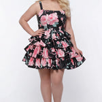 Plus-size model wearing a black and pink floral sleeveless mini dress with layered ruffle skirt. The dress features a square neckline, fitted bodice, and tiered ruffles at the waist and hem. She is styled with long wavy blonde hair, statement crystal earrings, and nude platform ankle-strap heels, posing against a studio background. Front view. 

Sydney's Closet SC8174
