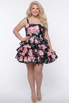 Plus-size model wearing a black and pink floral sleeveless mini dress with layered ruffle skirt. The dress features a square neckline, fitted bodice, and tiered ruffles at the waist and hem. She is styled with long wavy blonde hair, statement crystal earrings, and nude platform ankle-strap heels, posing against a studio background. Front view. 

Sydney's Closet SC8174