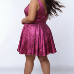 Plus-size model wearing a sleeveless a-line mini dress in a vibrant metallic fuchsia pink knit fabric. The dress features a deep V-neckline, fitted waistband, and a full flared skirt with subtle pleating. She is styled with long wavy dark brown hair, hoop earrings, and nude platform ankle-strap heels, posing against a light gray studio background.

Sydney's Closet SC8175 in magenta
