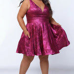 Plus-size model wearing a sleeveless a-line mini dress in a vibrant metallic fuchsia pink knit fabric. The dress features a deep V-neckline, fitted waistband, and a full flared skirt with subtle pleating. She is styled with long wavy dark brown hair, hoop earrings, and nude platform ankle-strap heels, posing against a light gray studio background while showing off the pockets in the skirt.

Sydney's Closet SC8175 in magenta