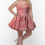 Plus-size model wearing a strapless a-line mini dress in pink floral brocade with metallic gold accents. The dress features a structured corset-style bodice with a strapless sweetheart neckline and a voluminous bubble hem skirt. She is styled with long wavy blonde hair, statement pink crystal earrings, and nude platform ankle-strap heels, posing against a light gray studio background.

Sydney's Closet SC8177 in pink floral