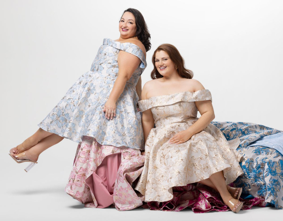 Two women wearing elegant off-shoulder dresses, playfully sitting on a couch covered in floral dresses.