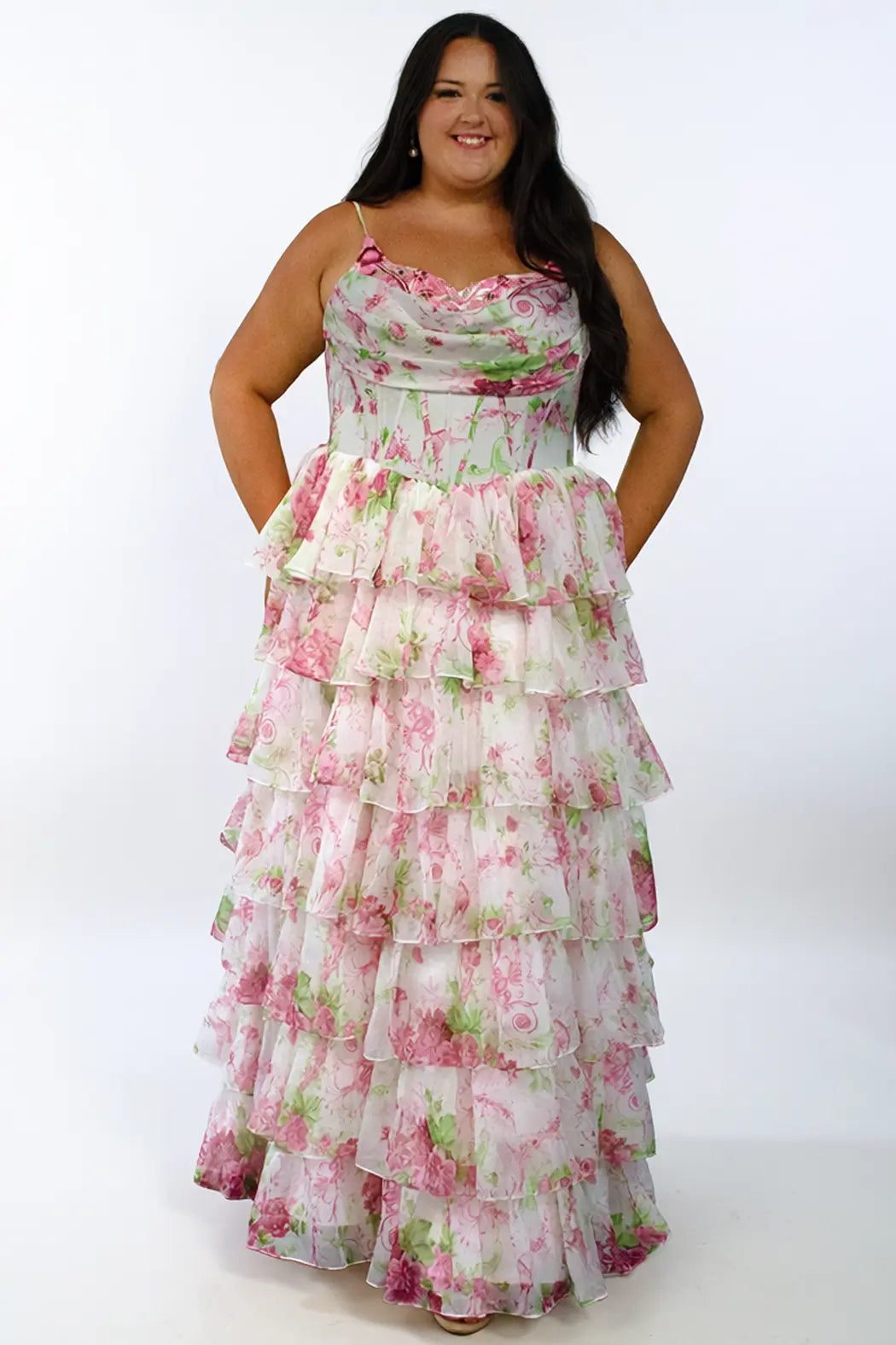 A teen modeling a sleeveless, tiered long prom dress with a pink and green floral print on a white background, photographed against a plain white studio backdrop. The dress features beaded embroidered appliques on the upper bust, pleated drape bodice, exposed boning, basque waistline and tiered chiffon skirt with slit. 

TE2702 in pink floral by Tease Prom, exclusively plus size prom dresses designed by Sydney's Closet