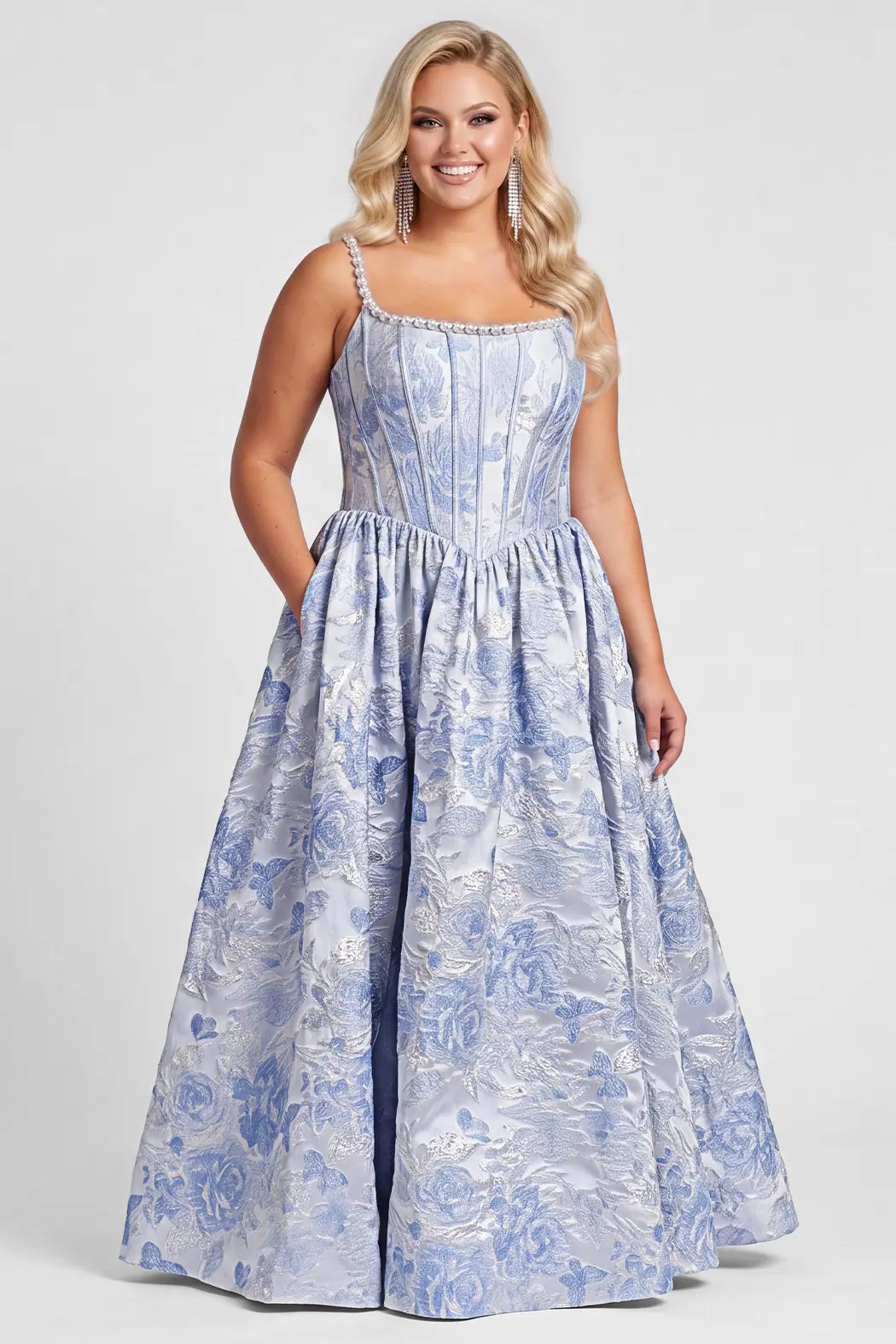 A blonde teen modeling a floor-length light blue and white floral brocade jacquard gown with pearl details on the stras and upper bodice, a structured corset-style bodice and a full A-line skirt, photographed against a plain light gray studio background.

TE2703 in blue floral by Tease Prom, exclusively plus size prom dresses designed by Sydney's Closet