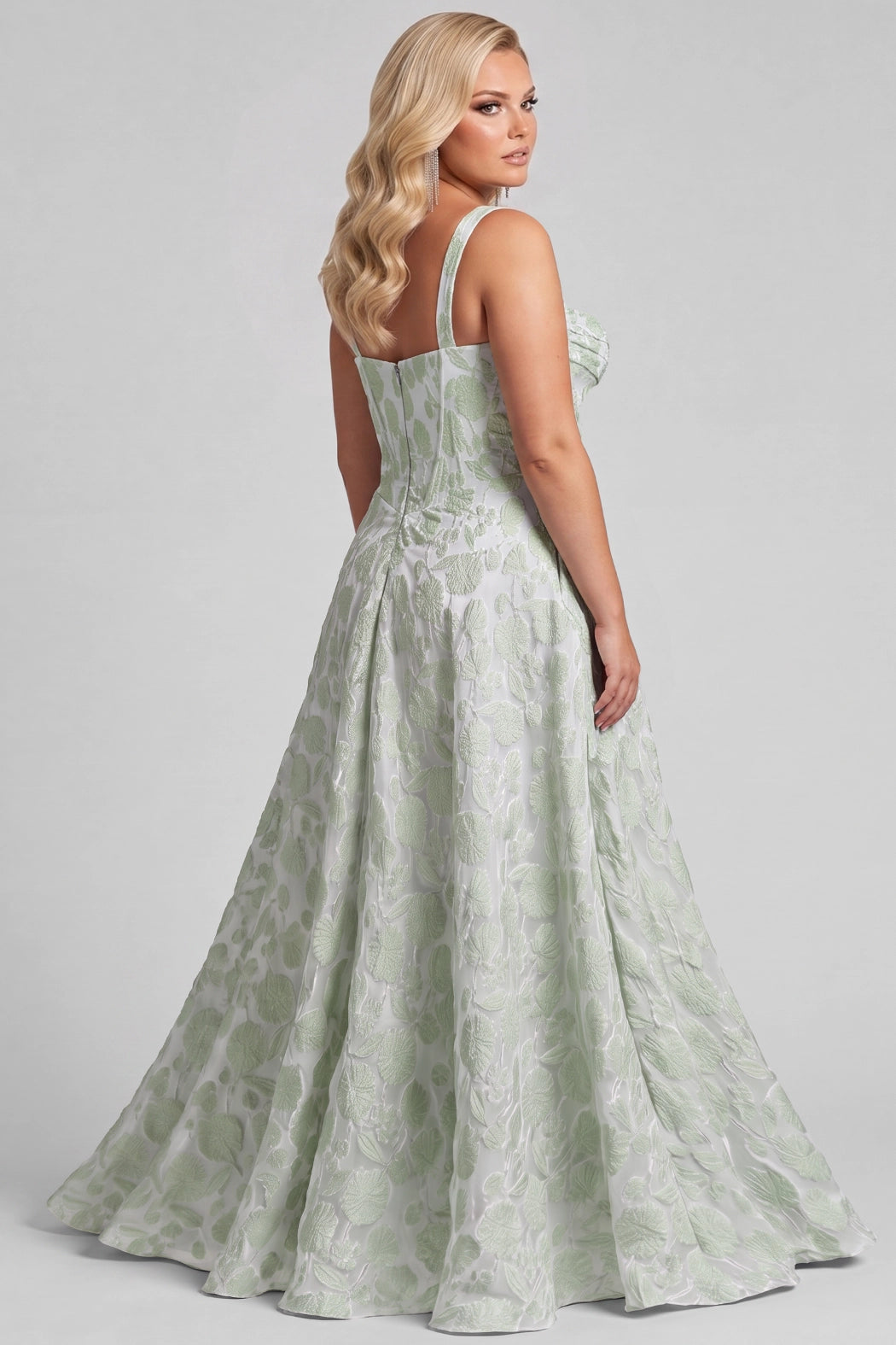 A blonde teen modeling a floor-length light green and brocade patterned gown with thin straps, a pleated bodice, and a high front slit, photographed against a plain light gray studio background.

TE2710 in sage green by Tease Prom, exclusively plus size prom dresses designed by Sydney's Closet.