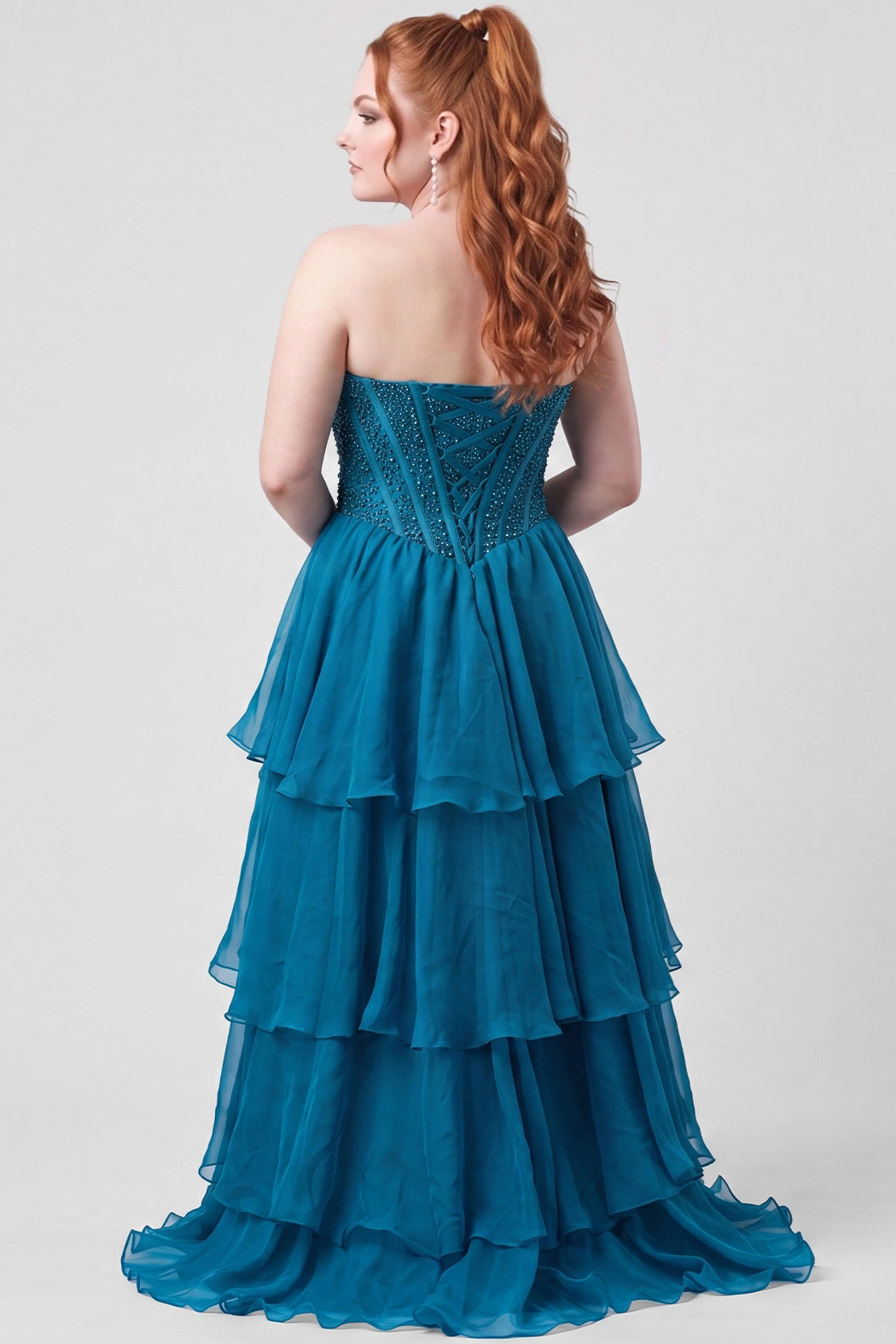 A red haired teen modeling a strapless teal blue gown with a beaded corset bodice and a tiered ruffled chiffon skirt with high slit, standing against a light gray studio background.

TE2717 in turquoise by Tease Prom, exclusively plus size prom dresses designed by Sydney's Closet