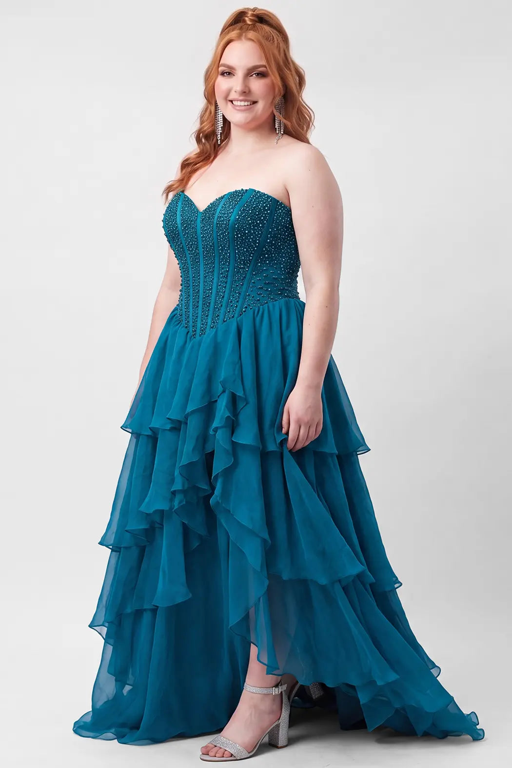 A red haired teen modeling a strapless teal blue gown with a beaded corset bodice and a tiered ruffled chiffon skirt with high slit, standing against a light gray studio background.

TE2717 in turquoise by Tease Prom, exclusively plus size prom dresses designed by Sydney's Closet