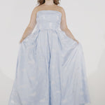 Woman models a strapless light blue A-line gown with a subtle floral pattern, standing in frame while playing with the skirt and placing her hands in the pockets.