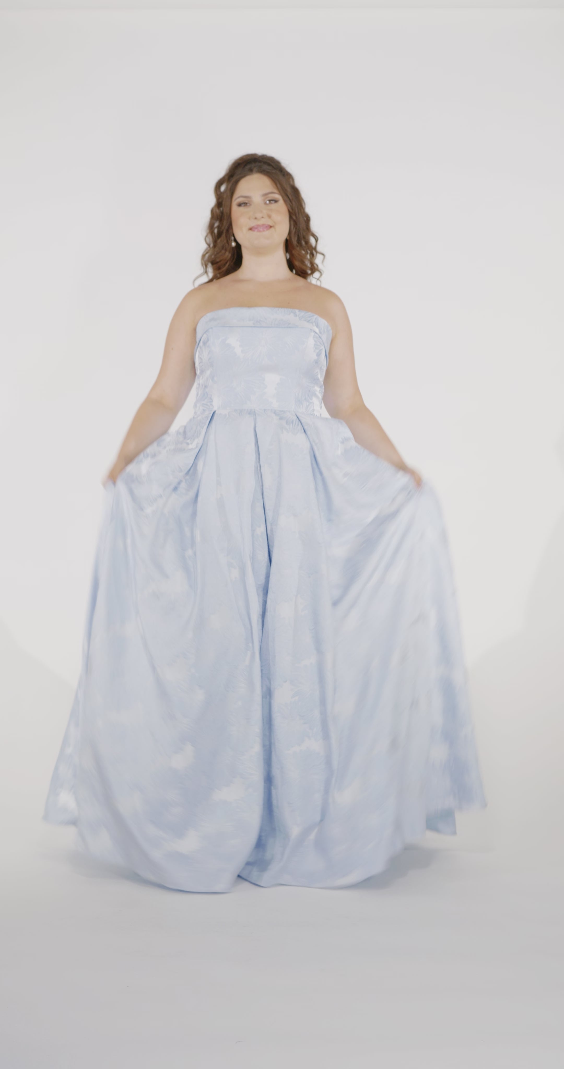Woman models a strapless light blue A-line gown with a subtle floral pattern, standing in frame while playing with the skirt and placing her hands in the pockets.
