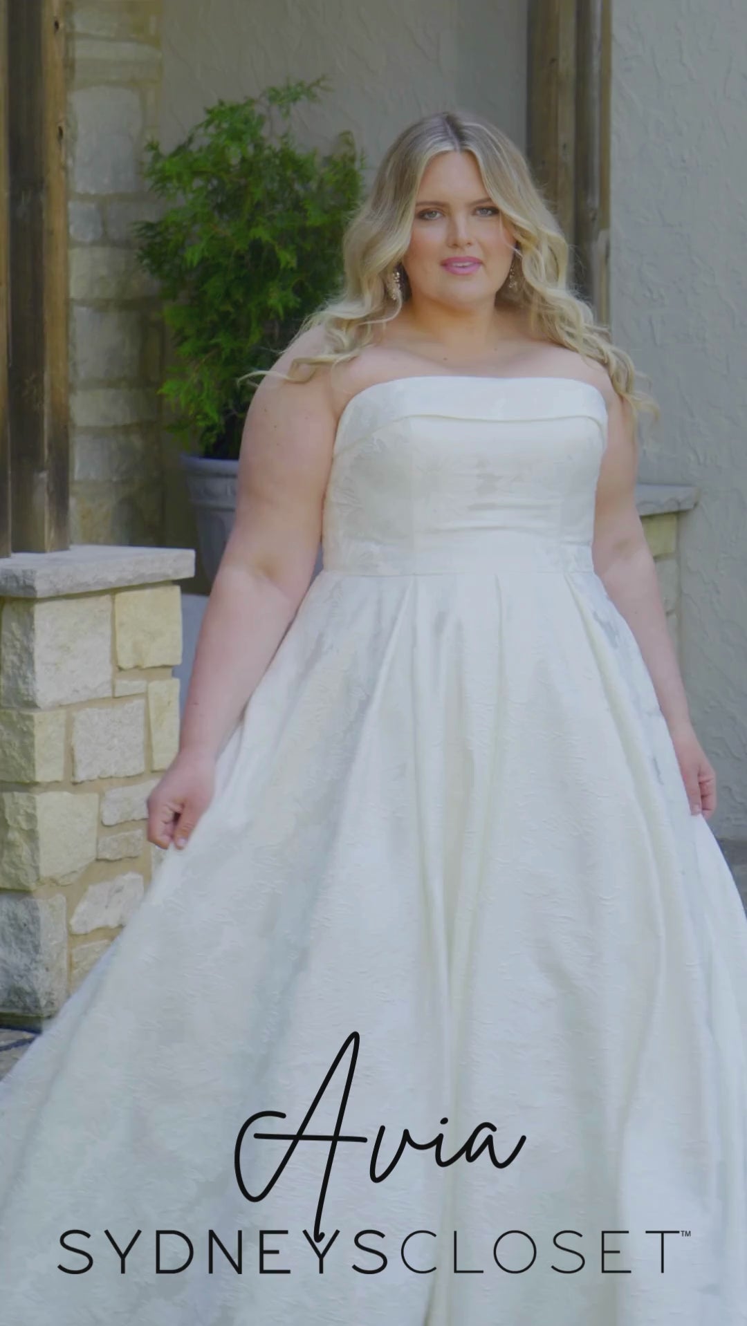 Video montage of a bride modeling a strapless A-line wedding gown with a subtle floral print, shown outdoors from multiple angles and distances. The text “Avia” and the “Sydney's Closet” logo appear at the bottom.