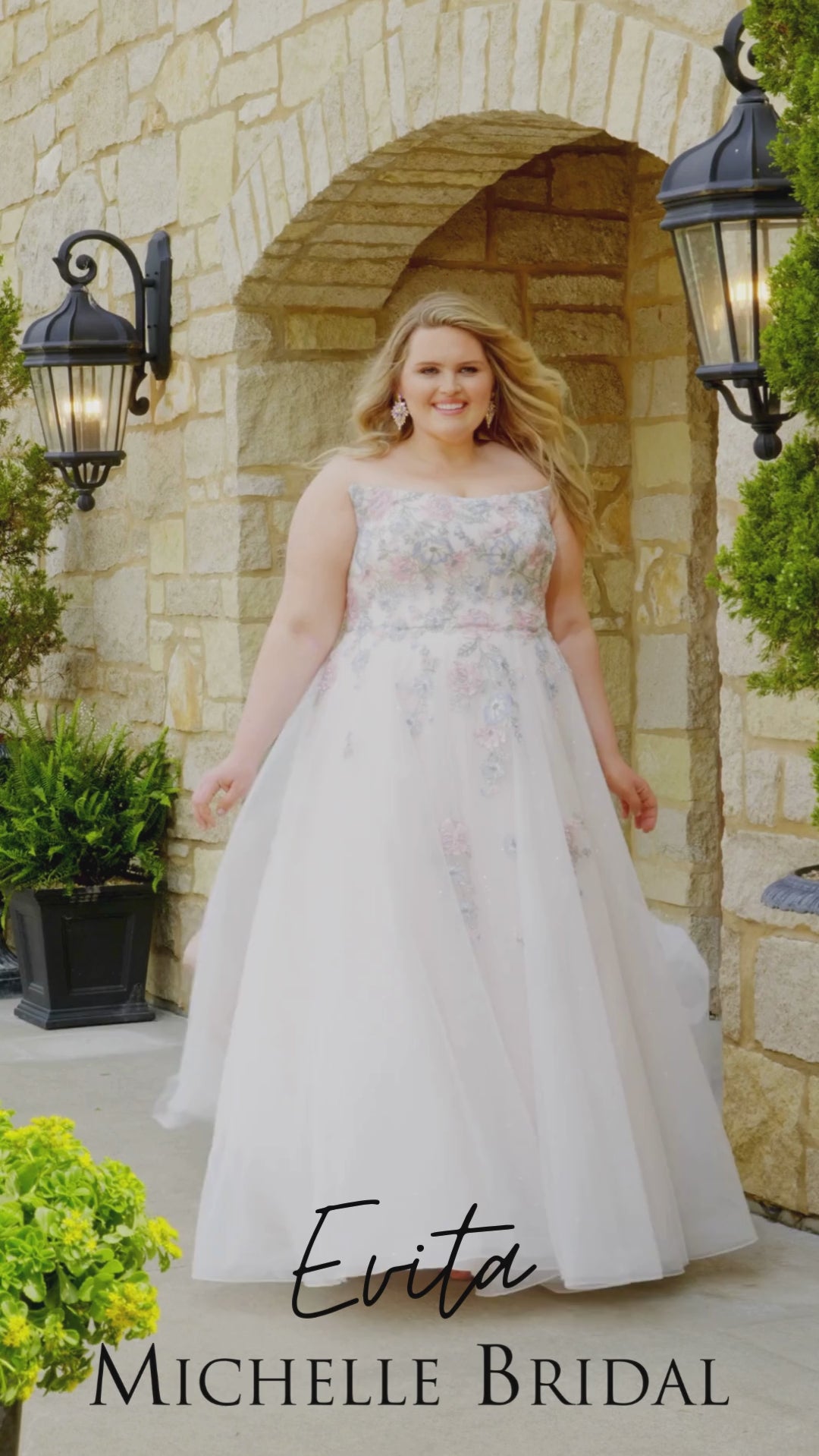Plus-size bride wearing the Michelle Bridal MB2614 Evita wedding gown with pastel blue and pink embroidered lace appliqués on the bodice, a strapless scoop neckline, and a sparkle tulle A-line skirt.