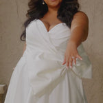 Video montage of a bride modeling a strapless white A-line wedding gown with a sweetheart neckline and large bow on the waistline, shown outdoors from multiple angles and distances. The text “Saphira” and the “Sydney's Closet” logo appear at the bottom.