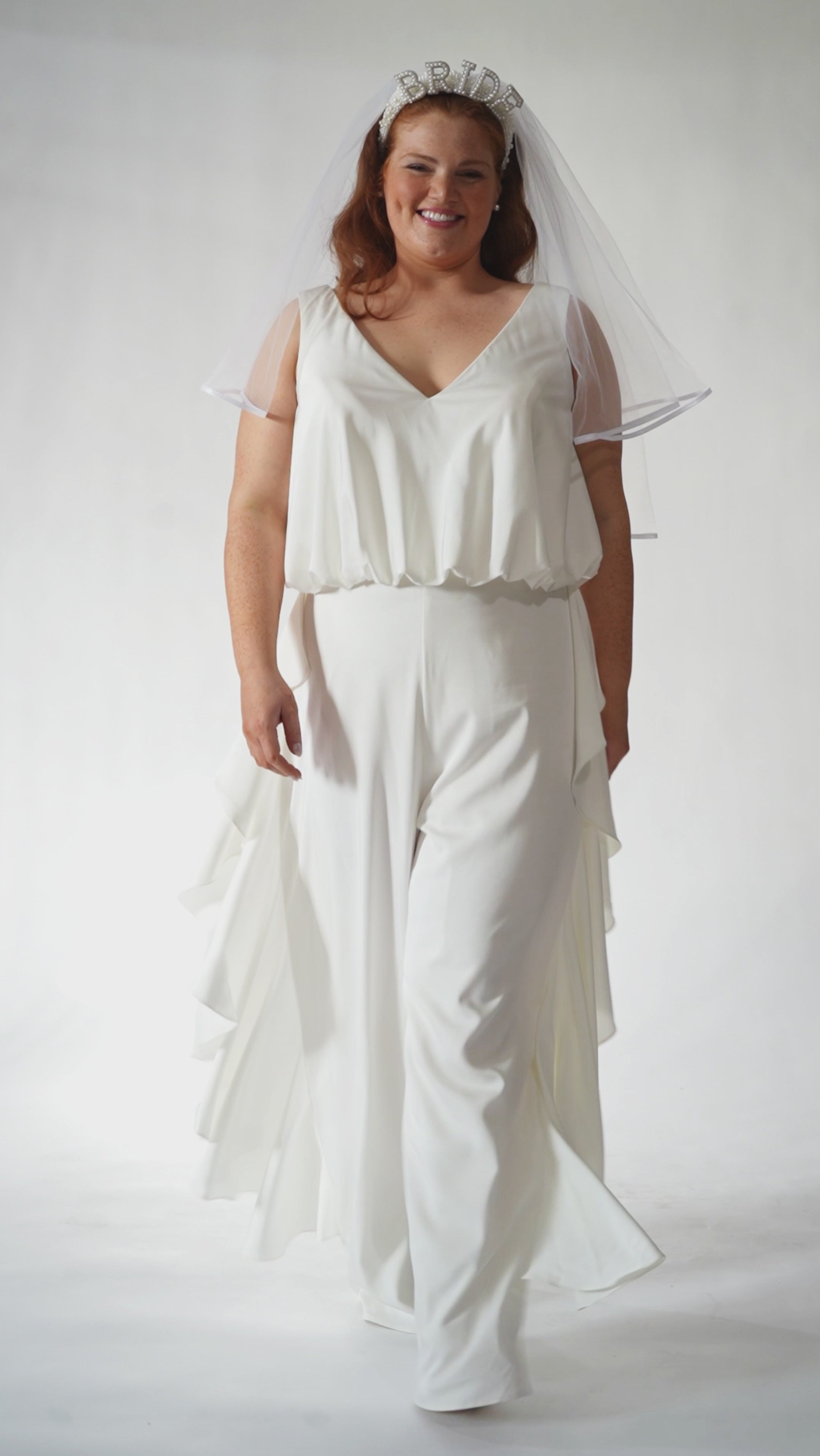 Celebration's by Sydney's Closet style CE2615 in Ivory. 

Crepe satin jumpsuit with a V-neck, bra-friendly straps, wide leg pants and blouson bodice. Ruffle detail on outer seams of pants. V-back with long center back zipper. 