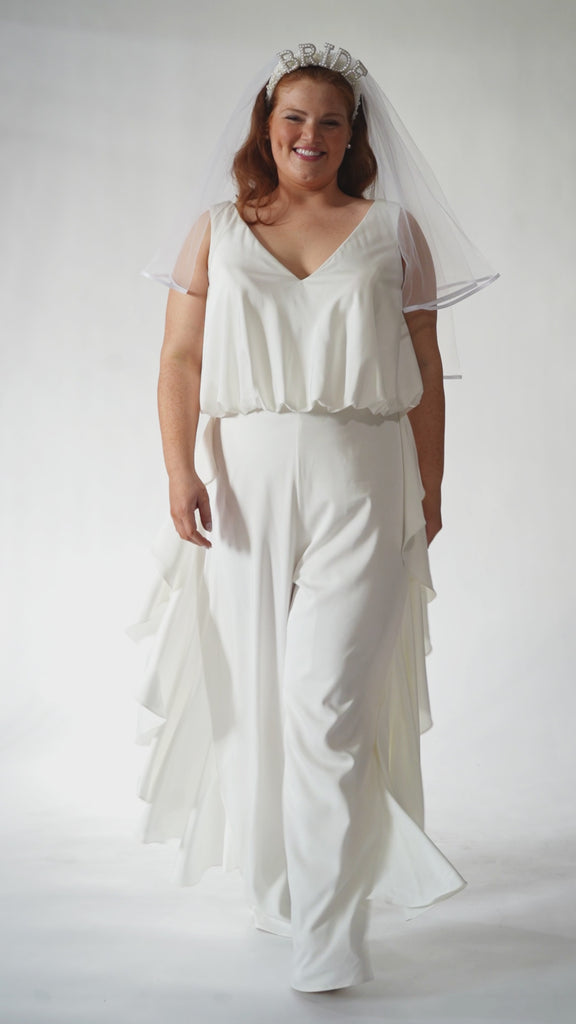 Celebration's by Sydney's Closet style CE2615 in Ivory. 

Crepe satin jumpsuit with a V-neck, bra-friendly straps, wide leg pants and blouson bodice. Ruffle detail on outer seams of pants. V-back with long center back zipper. 