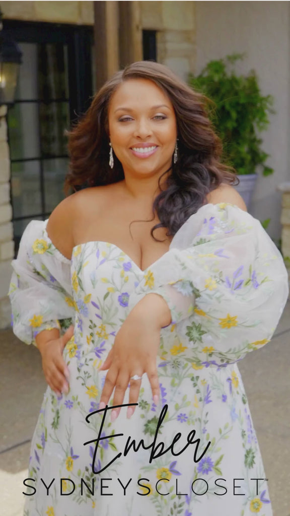 Video montage of a woman modeling a strapless A-line gown with sheer puff sleeves and colorful floral embroidery, shown outdoors from multiple angles and distances. The text “Ember” and the “Sydney's Closet” logo appear at the bottom.