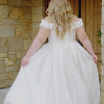 Plus-size bride in the Michelle Bridal MB2610 Marcella wedding gown in ivory Mikado satin, featuring an off-the-shoulder sweetheart neckline, pearl and sequin accents, and a box-pleated A-line skirt.