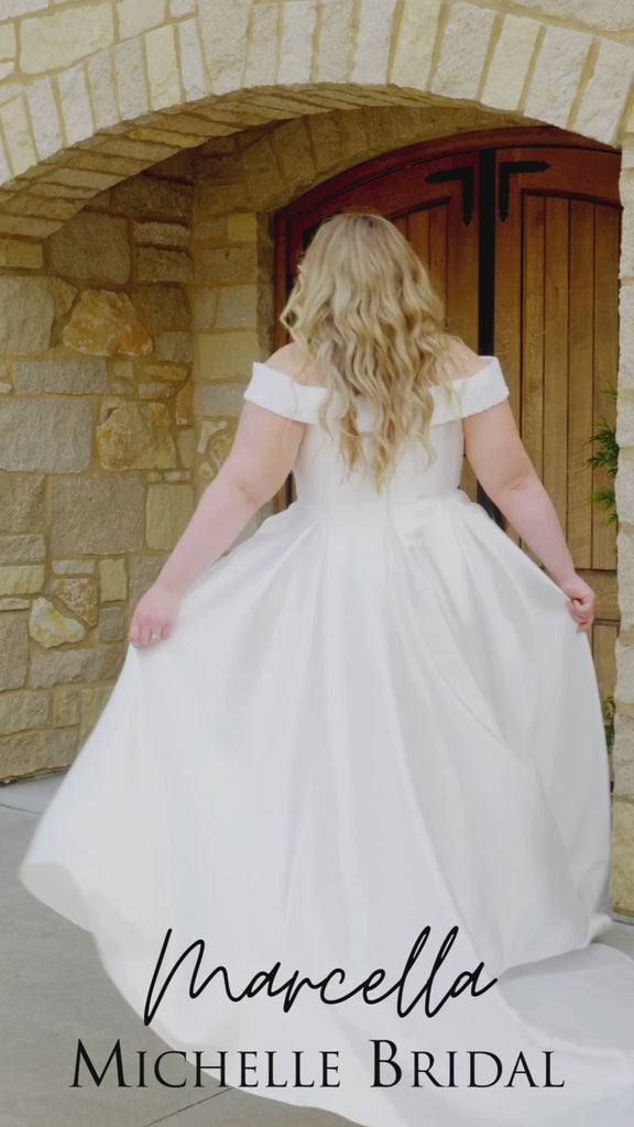 Plus-size bride in the Michelle Bridal MB2610 Marcella wedding gown in ivory Mikado satin, featuring an off-the-shoulder sweetheart neckline, pearl and sequin accents, and a box-pleated A-line skirt.
